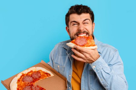 Foto de Hombre divertido comiendo pizza de pie sosteniendo la caja de pizzería sobre fondo azul en el estudio. Tipo hambriento disfrutando de la comida chatarra, saboreando sabrosa comida poco saludable. Malos hábitos nutricionales, concepto de entrega - Imagen libre de derechos