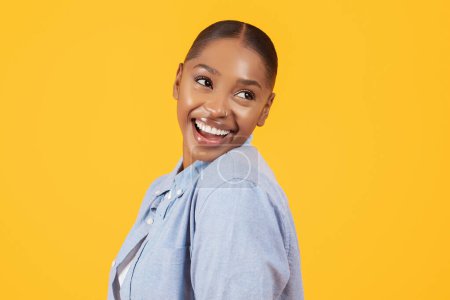 Téléchargez les photos : Portrait de jolie femme noire ludique en chemise denim décontractée, souriant regardant de côté tout en posant sur fond de studio jaune. Joyeux modèle de dame afro-américaine pose pour photo - en image libre de droit