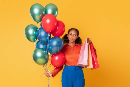 Téléchargez les photos : Ventes de vacances. Adolescente africaine américaine réfléchie tient des sacs à provisions en papier et un tas de ballons colorés debout sur fond de studio jaune, pensant à de grandes offres de commerce festif - en image libre de droit