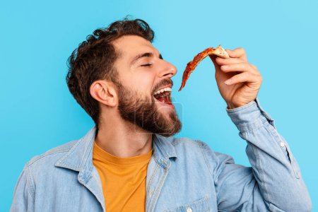 Photo for Fast Food. Closeup Shot Of Hungry Guy Eating Pizza Holding Slice Near Opened Mouth, Enjoying Junk Food Over Blue Background In Studio. Binge Eating And Unhealthy Nutrition Habits - Royalty Free Image