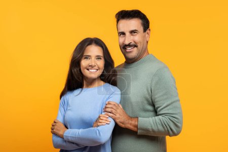 Foto de Una feliz pareja caucásica feliz se para de cerca, con el brazo del hombre alrededor de la mujer, ambos sonriendo con confianza hacia la cámara contra un alegre fondo naranja - Imagen libre de derechos