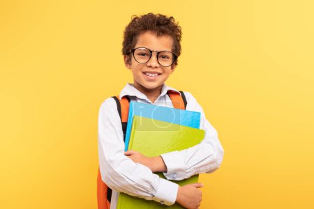 Foto de Joven estudiante confiado con pelo rizado y gafas, sosteniendo carpetas de colores, usando camisa blanca con tirantes, mochila naranja contra fondo amarillo, listo para la escuela - Imagen libre de derechos