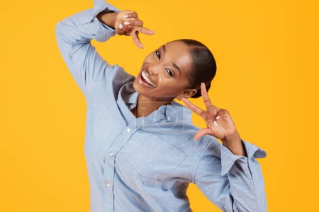 Téléchargez les photos : Portrait de femme afro-américaine positive montrant des symboles de signe V près du visage, s'amuser et tromper sur fond jaune, porter une chemise en denim décontractée et sourire à la caméra - en image libre de droit