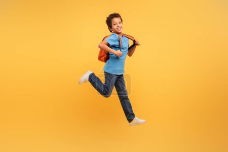 Photo for Energetic young boy with curly hair leaping joyfully, wearing blue shirt and backpack, against dynamic yellow background, capturing the essence of carefree childhood fun - Royalty Free Image