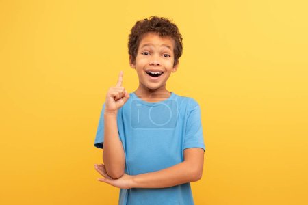 Téléchargez les photos : Jeune garçon excité aux cheveux bouclés, les yeux grands ouverts d'excitation, levant le doigt dans le moment eureka classique, portant une chemise bleue sur fond jaune, suggérant créativité et perspicacité - en image libre de droit