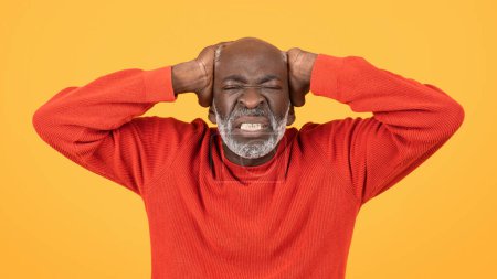 Photo for Frustrated sad senior man in a red sweater and hat, clutching his head with a grimace, standing against a yellow background, conveying strong emotion and stress, close up, panorama - Royalty Free Image