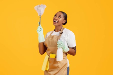 Foto de Limpiador de mujeres afroamericanas felices limpiando y espolvoreando con plumas, sosteniendo aerosol de detergente, disfrutando de la limpieza de primavera sobre el fondo del estudio amarillo, usando delantal - Imagen libre de derechos
