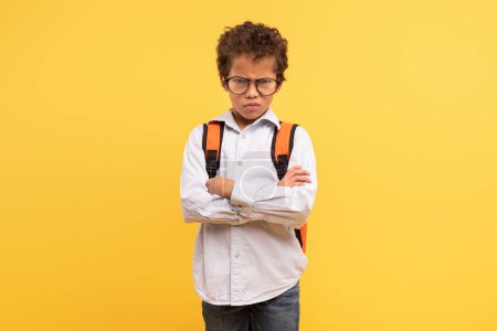Photo for Angry schoolboy with curly hair and eyeglasses, arms crossed in questioning stance, wearing shirt and backpack against yellow background, embodying skepticism or critical thinking - Royalty Free Image