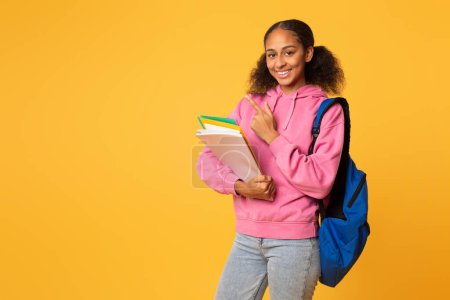 Téléchargez les photos : Joyeux étudiant afro-américain tenant des classeurs et des cahiers d'université, posant avec un sac à dos bleu et pointant du doigt vers l'espace libre sur fond de studio jaune. Offre éducative - en image libre de droit