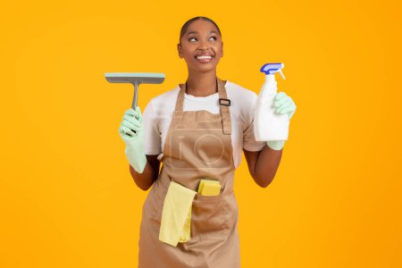 Téléchargez les photos : Femme de ménage noire souriante avec spray nettoyant et brosse à raclette pour fenêtres, se préparant au nettoyage du printemps, exsudant de la positivité dans son travail sur fond de studio jaune. Concept de service de ménage - en image libre de droit
