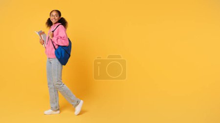 Téléchargez les photos : Offre éducative. Étudiante adolescente afro-américaine tient des classeurs d'université posant avec son sac à dos bleu, debout près de l'espace vide pour le texte sur fond de studio jaune. Panorama - en image libre de droit