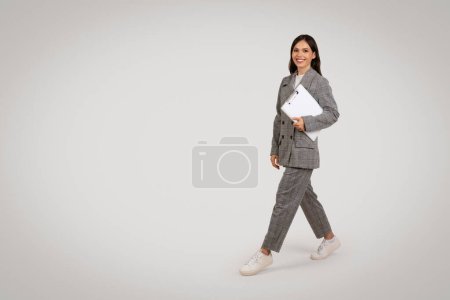 Foto de Dinámico joven profesional con confianza caminando con el ordenador portátil en la mano, vestido con traje a cuadros de moda emparejado con zapatillas de deporte casuales, que encarna traje casual de negocios moderno, longitud completa - Imagen libre de derechos