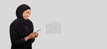 Téléchargez les photos : Femme d'affaires afro-américaine souriante dans un hijab engagé dans une conversation agréable sur son téléphone portable, rayonnant de positivité et de confiance professionnelle sur fond gris, studio - en image libre de droit