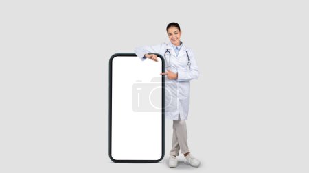 Foto de Un joven médico europeo alegre en una bata de laboratorio apunta a una pantalla de teléfono inteligente en blanco, perfecta para mostrar aplicaciones de salud digitales, contra un fondo de estudio gris - Imagen libre de derechos