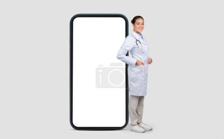 Foto de Una doctora europea sonriente en una bata blanca de laboratorio con un estetoscopio está de pie con confianza junto a un teléfono inteligente de gran tamaño, que simboliza la telemedicina y los servicios de salud modernos - Imagen libre de derechos