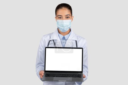 Téléchargez les photos : Médecin européen professionnel en blouse blanche et masque protecteur, présentant un ordinateur portable ouvert avec un écran vierge pour l'affichage de l'information médicale, isolé sur fond de studio gris - en image libre de droit