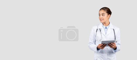 Foto de Una doctora europea sonriente con una bata de laboratorio y un estetoscopio mira a un lado mientras sostiene una tableta, lo que representa la integración de la tecnología en la atención al paciente y la gestión de los registros médicos. - Imagen libre de derechos