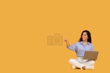 Téléchargez les photos : Joyeux jeune femme dans un chemisier bleu et pantalon blanc assis jambes croisées sur le sol, tenant un ordinateur portable sur ses genoux et pointant vers le haut avec un sourire, indiquant une idée créative ou une découverte - en image libre de droit
