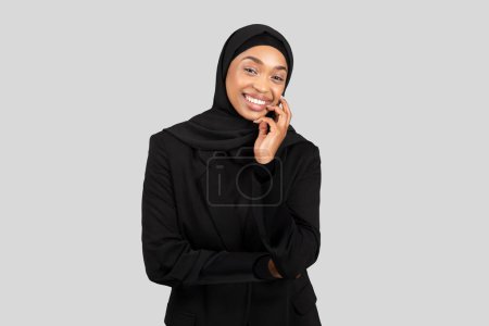 Foto de Mujer de negocios afroamericana sonriente en un hiyab que participa en una conversación telefónica, ejemplificando la comunicación profesional en un entorno corporativo multicultural sobre fondo gris, estudio - Imagen libre de derechos