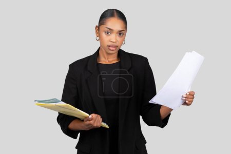 Téléchargez les photos : Femme d'affaires afro-américaine sérieuse tenant des documents et un dossier, à la recherche d'une réunion ou d'un audit important, dans un costume noir professionnel sur fond gris, studio - en image libre de droit