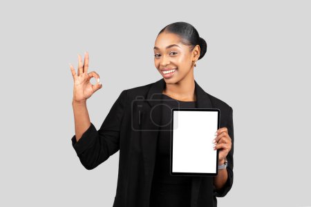 Foto de Empresaria afroamericana positiva sosteniendo una tableta con una pantalla en blanco mientras hace un gesto OK, simbolizando la satisfacción con las soluciones digitales, en un traje negro - Imagen libre de derechos