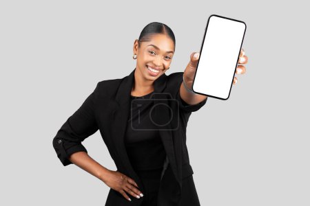 Téléchargez les photos : Femme d'affaires afro-américaine joyeuse présentant un écran de smartphone, prête à démontrer une nouvelle application ou un site Web mobile, portant un costume noir professionnel sur fond gris, studio - en image libre de droit