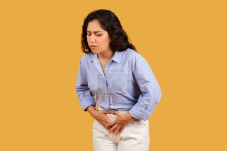 Photo for Woman with a pained expression, clutching her abdomen, possibly experiencing stomach pain or discomfort, dressed in a blue shirt and cream pants against a yellow background - Royalty Free Image