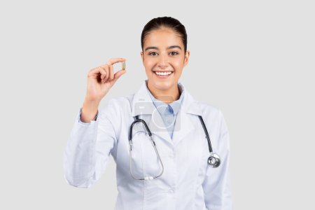 Photo for Happy european doctor in a white lab coat holding up a single capsule with a stethoscope around her neck, symbolizing medical treatment and healthcare services, isolated on gray studio background - Royalty Free Image