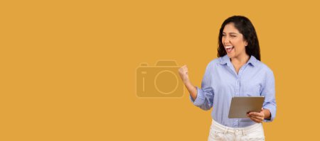 Foto de Mujer joven exuberante con el pelo rizado riendo y celebrando el éxito mientras sostiene una tableta, mostrando un gesto de puño bomba, vestido con una camisa azul claro sobre un fondo naranja - Imagen libre de derechos