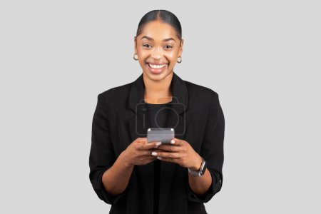 Téléchargez les photos : Femme d'affaires afro-américaine souriante utilisant un smartphone, engagée dans la communication moderne, représentant l'accessibilité et la connectivité dans une tenue professionnelle sur fond gris, studio - en image libre de droit