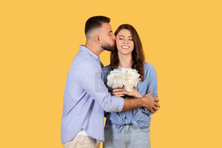 Téléchargez les photos : Moment délicat alors que l'homme embrasse sa partenaire sur la joue, tandis qu'elle ferme les yeux et sourit, tenant un bouquet de fleurs blanches, sur fond jaune - en image libre de droit
