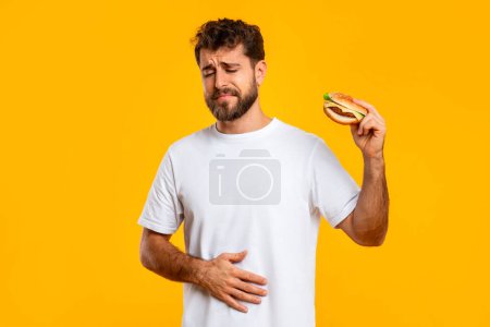 Téléchargez les photos : Manger de façon excessive malsaine. Homme bouleversé touchant l'estomac douloureux tenant un hamburger, souffrant de maux d'estomac après avoir mangé de la malbouffe malsaine sur fond jaune studio. Douleur abdominale - en image libre de droit