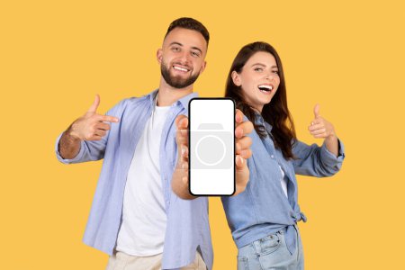 Foto de Hombre y mujer alegre dando pulgares hacia arriba y apuntando a la pantalla del teléfono inteligente en blanco, ideal para la promoción de aplicaciones, contra el fondo amarillo soleado - Imagen libre de derechos