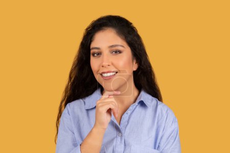 Foto de Una joven confiada con el pelo oscuro ondulado y una sonrisa radiante, posando pensativamente con la mano en la barbilla, llevando una camisa azul casual sobre un fondo amarillo vibrante - Imagen libre de derechos