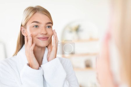 Téléchargez les photos : Femme blonde sourit tout en touchant le visage et en regardant le miroir, profitant de sa peau douce parfaite après le traitement de soins de la peau dans un cadre de salle de bain moderne. Focus sélectif, espace libre - en image libre de droit