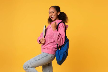 Photo for Joyful african american teenager student girl gesturing yes holding college books and wearing backpack, over yellow background, celebrating academic success at college. Studio shot - Royalty Free Image