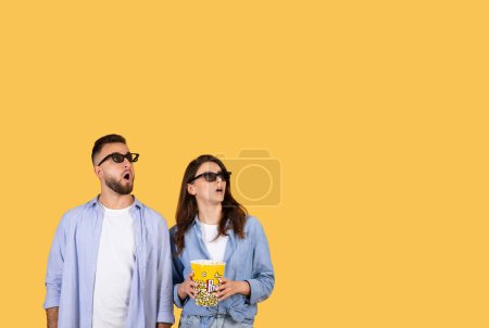 Foto de Hombre y mujer en gafas 3D miran hacia arriba en el espacio libre con asombro, sosteniendo palomitas de maíz cubo, lo que sugiere una experiencia de película inmersiva, contra fondo amarillo vivo - Imagen libre de derechos