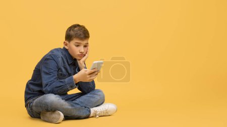 Foto de Niño adolescente aburrido usando un teléfono inteligente mientras está sentado en el piso en el estudio, niño varón molesto mirando la pantalla del teléfono móvil, jugando juegos en línea, sentirse solo, posando sobre un fondo amarillo, espacio para copiar - Imagen libre de derechos