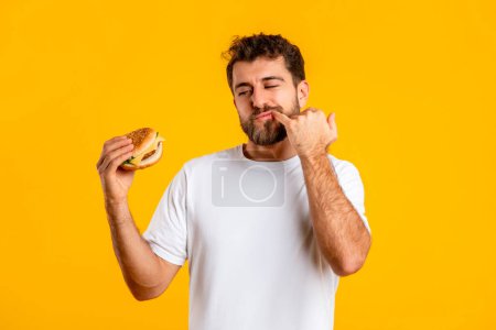 Téléchargez les photos : Plaisir coupable. Portrait d'un homme drôle affamé tenant un hamburger à la main, mangeant de la restauration rapide malsaine, sur fond de studio jaune. Un affamé excité veut un cheeseburger qui lèche les doigts. Délicieux en-cas - en image libre de droit