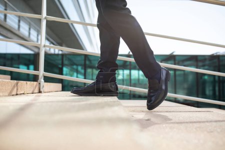 Photo for Cropped shot of male legs as unrecognizable businessman walking upstairs, taking confident steps on city stairs, symbolizing career opportunities and growth, in urban area outside - Royalty Free Image