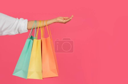 Téléchargez les photos : Sacs à provisions colorés suspendus à la main féminine sur fond de studio rose, femme commerçante méconnaissable portant des achats, bénéficiant de ventes saisonnières et de réductions, plan recadré avec espace de copie - en image libre de droit