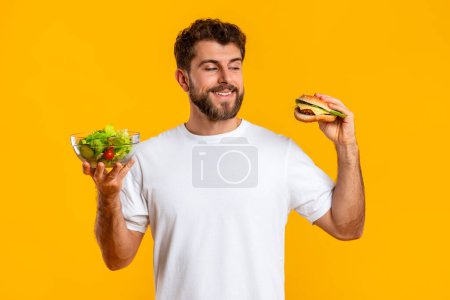 Téléchargez les photos : Vs en bonne santé Malsain. Jeune homme affamé Choisir entre Burger et Salade Repas posant debout sur fond jaune Studio. Nutrition et choix d'aliments, concept de repas de cheat sur un régime alimentaire - en image libre de droit