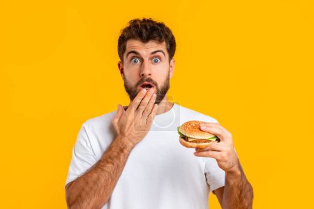 Photo for Oops, Cheat Meal. Portrait Of Funny Shocked Young Guy Eating Tasty Burger During Diet, Posing On Yellow Background In Studio. Junk Food Is My Guilty Pleasure, Concept Of Unhealthy Food - Royalty Free Image