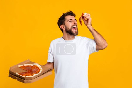 Photo for Hungry Millennial Man Eating Pizza Standing And Holding Pizzeria Box Over Yellow Studio Backdrop. Portrait Shot Of Guy In White T-Shirt Savoring Fastfood. Tasty Junk Food, Pizza Delivery Concept - Royalty Free Image