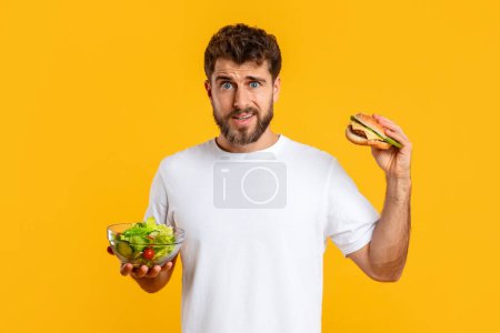 Photo for Food Choice Problem. Hungry Confused Guy Holding Burger And Fresh Vegetable Salad Choosing Diet Over Yellow Studio Background. Healthy Vs Unhealthy Nutrition, Junk Food Concept - Royalty Free Image