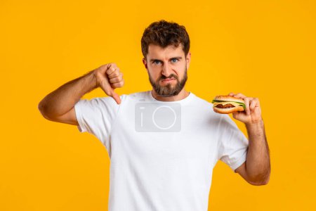 Photo for Dont Eat Junk Food. Young Man Holding Burger And Gesturing Thumb Down, Disapproving Unhealthy Bad Nutrition Habit With Disgust, Standing On Yellow Studio Background. Fastfood, Dislike - Royalty Free Image