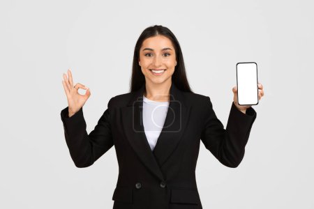 Photo for Glad caucaisan woman in formal suit showing smartphone with empty screen and ok sign with hand, isolated on gray studio background. Business recommendation, app, website - Royalty Free Image