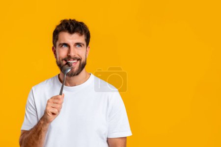 Photo for Hungry guy holding and biting spoon looking at free space aside, posing on yellow studio background. Portrait of funny young man advertising food offer, waiting for dinner. Male nutrition concept - Royalty Free Image