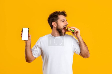Photo for Hungry young man with burger shows smartphone blank screen eating over yellow studio background. Guy recommending mobile application for junk food delivery or weight loss app. Mockup - Royalty Free Image
