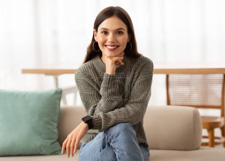 Photo for Attractive young brunette woman wearing casual clothing sitting on couch at home, showing her beautiful cozy living room interior, smiling at camera, copy space - Royalty Free Image